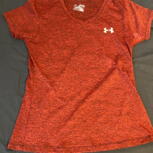 Röd Under Armour t-shirt loose fit - Storlek M men passar även S. Snygg rödmelerad t-shirt från Under Armour med v-ringning och korta ärmar. T-shirten har vit logga på bröstet och är i loose fit för en avslappnad känsla. Perfekt för träning eller chill dagar. Tillverkad i funktionsmaterial som andas.