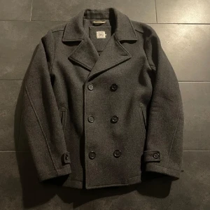 Mörkgrå skepparkavaj i ull - Stilren mörkgrå skepparkavaj med dubbelknäppning och klassisk krage. Jackan har två snedställda fickor fram och snygga knapprade detaljer vid ärmsluten. Perfekt för dig som vill ha en tidlös och clean look under kyliga dagar.