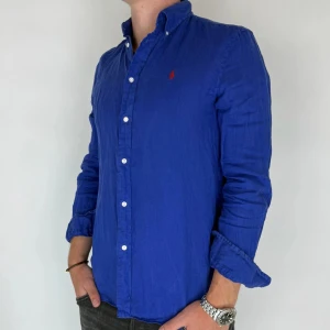 Ralph Lauren Linne Skjorta - Nu har vi nöjet att erbjuda en klassisk skjorta från det välkända varumärket Ralph Lauren. Plagget är i storlek Medium och är i mycket gott skick, utan några synliga defekter. Modellen på bilden är 180 cm lång och väger 75 kg, vilket kan ge en god indikation på tröjans passform.