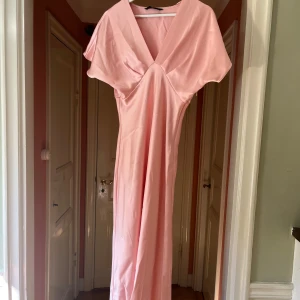 Ljusrosa långklänning från Zara - Supersnygg ljusrosa långklänning från Zara i satinliknande tyg. Klänningen har v-ringning, korta ärmar och en elegant, figurnära passform. Perfekt för dig som vill sticka ut med en feminin och stilren look.