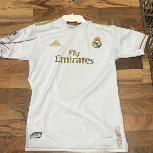 Real Madrid Benzema 9 Adidas tröja - Vit Real Madrid matchtröja med guldiga detaljer, Adidas-logga och Fly Emirates-tryck på bröstet. Benzema och nummer 9 på ryggen. La Liga-märke på ärmen. Tillverkad i lätt och ventilerande material (Climacool). Klassisk passform med korta ärmar.