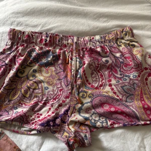 Färgglada paisley shorts från Primark - Snygga shorts från Primark i storlek M med färgstarkt paisleymönster i rosa, lila, gult och blått. De har elastisk midja med snörning och är gjorda i ett mjukt, lätt material som känns skönt mot huden. Perfekta för varma dagar när du vill sticka ut lite extra.