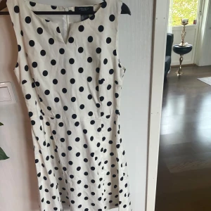Vit prickig klänning från Sara Kelly - Supersöt vit klänning med svarta prickar från Sara Kelly. Ärmlös modell med figurnära passform och diskret v-ringning framtill. Dragkedja i ryggen för enkel påklädning. 