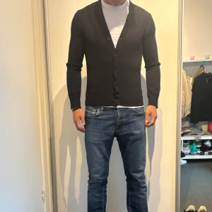 Svart kofta med knappar - En stilren mörkblå kofta med djup v-ringning och knäppning framtill. Modellen är långärmad och har en slim passform som sitter snyggt på kroppen. Perfekt att slänga över en t-shirt för en clean look. Materialet känns mjukt och stretchigt och är i merinoull. Använd 5 gånger. Köpt på brothers för 1300 kr. 