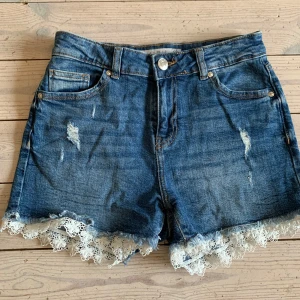 Jeansshorts med spets från New Yorker - Blå jeansshorts från New Yorker med snygga slitningar och vit spetskant nertill. Klassisk femficksmodell med knapp och dragkedja framtill. Perfekta för en chill sommardag, med en touch av vintagekänsla tack vare spetsdetaljerna.