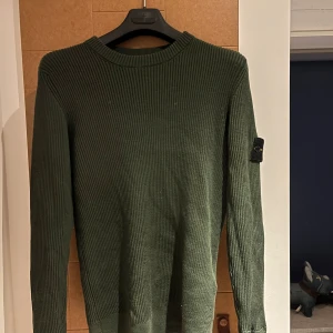 Grön stickad tröja från Stone Island - Säljer en grön ribbstickad tröja från Stone Island med klassisk patch på ärmen. Tröjan har rund halsringning och långa ärmar, perfekt för lager-på-lager under höst och vinter. Materialet är mjukt och skönt, och passformen är normal.