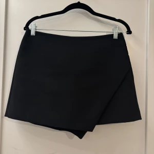 Svart omlott kortkjol - Stilren svart kortkjol med omlott-design framtill och inbyggda shorts från Zara. Använd endast en gång. Jag anser att kjolen är liten i storleken och borde passa någon som brukar bära storlek M.