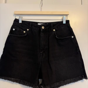 Svarta jeansshorts från NA-KD - Snygga svarta jeansshorts från NA-KD med hög midja och råa fransade benslut. Klassisk femficksmodell med knapp och dragkedja framtill. Perfekta för en cool och avslappnad stil under sommaren.