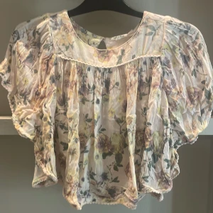 Blommig blus från KappAhl - Supersöt blommig blus från KappAhl i ljusa pastellfärger. Modellen är croppad med volangiga, korta ärmar och rynkade detaljer. Tunt, luftigt tyg som känns perfekt för varmare dagar. Blusen har en romantisk vibe med sitt blommönster och mjuka färgskala.