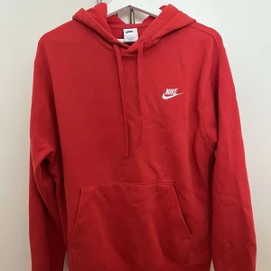 Röd Nike hoodie med huva - Röd hoodie från Nike med klassisk logga broderad i vitt på bröstet. Tröjan har huva med snörning och en stor känguruficka framtill. Perfekt för dig som gillar en sportig och avslappnad stil.