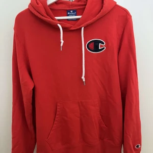 Röd hoodie från Champion med logga - Röd hoodie från Champion med stor broderad logga på bröstet och klassisk känguruficka. Tröjan har vit snörning i huvan och ribbade muddar. Perfekt för dig som gillar sportig och avslappnad stil.