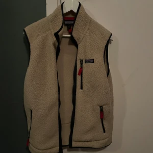 Beige/vit  fleeceväst från Patagonia - Säljer en beige fleeceväst från Patagonia med svarta detaljer och röda dragkedjor. Västen har hög krage, tre fickor med dragkedja och klassisk Patagonia-logga på bröstet. Perfekt att slänga över en hoodie eller t-shirt för extra värme.