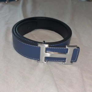 H blått skärp med H-spänne - H bälte som man kan vända håll och göra till svart om man vill.Snyggt blått skärp från H med ikoniskt H-spänne i silverfärgad metall. Bältet är gjort i slätt skinn med vita kontrastsömmar och har en stilren, klassisk design som sticker ut. Perfekt accessoar för att lyfta din outfit.