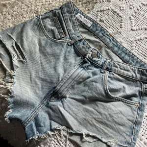 Ljusa jeansshorts med slitningar från Zara - Snygga ljusblå jeansshorts från Zara med råa, fransiga kanter och slitningar på ena benet. Klassisk femficksmodell med bälteshällor och dragkedja. Perfekta för en avslappnad och trendig look under varma dagar.