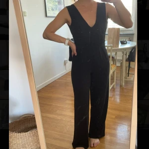 Svart ärmlös jumpsuit med v-ringning - Snygg svart ärmlös jumpsuit med djup v-ringning både fram och bak. Vida ben och markerad midja ger en stilren och modern look. Perfekt för dig som gillar minimalistisk stil och vill ha något bekvämt men ändå dressat.