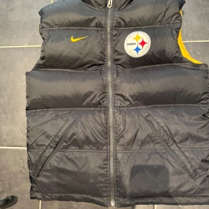 Reversibel Steelers dunväst från Nike - Reversibel dunväst från Nike med Pittsburgh Steelers-logga. Ena sidan är svart med Steelers-märke och gul detalj, andra sidan är helgul med svart Steelers-tryck och NFL Pro Line-märke. Västen har dragkedja och två fickor framtill. Perfekt för dig som vill sticka ut med sportig stil.