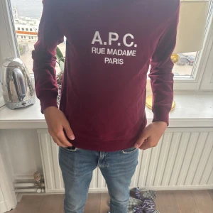 Vinröd sweatshirt från A.P.C. - Vinröd sweatshirt från A.P.C. med vit broderad text 'A.P.C. RUE MADAME PARIS' på bröstet. Klassisk rund hals, långa ärmar och ribbade muddar. Skön passform och tillverkad i mjuk bomull, perfekt för en clean och stilren look.