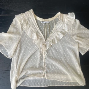Vit blus med volang från Zara - Supersöt vit blus från Zara med korta ärmar, volang längs v-ringningen och små prickar i tyget. Blusen är genomskinlig och har knappar. Perfekt för dig som gillar en feminin och trendig stil. Frakt ingår ej 