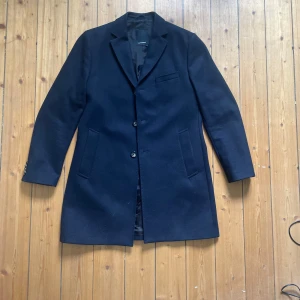 Mörkblå rock med klassisk krage - Snygg mörkblå rock från J.lindeberg med klassisk krage och knäppning framtill. Jackan har två fickor på sidorna och en bröstficka, samt slät design utan synliga detaljer. Helt oanvänd!