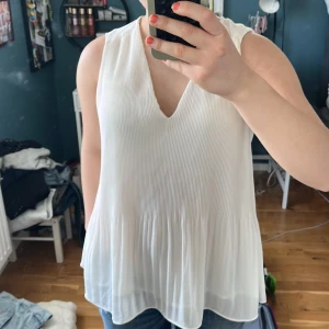 Vit plisserad ärmlös blus - Säljer en luftig vit blus med plisserat tyg och v-ringning. Toppen är ärmlös och har en flowy passform som ger en avslappnad men ändå stilren look. Perfekt att matcha med shorts eller kjol för en fräsch outfit.