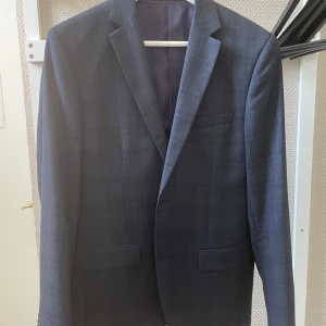 Blå rutig kavaj från H&M, slim fit - Snygg blå kavaj från H&M i slim fit-modell med diskret rutigt mönster. Tillverkad i en blandning av ull och polyester, fodrad med viskos och polyester. Klassisk design med två fickor fram och en bröstficka. Perfekt för dig som gillar stilrena detaljer.
