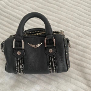 Svart Zadig väska - Cool svart handväska från Zadig i skinn med silvriga detaljer, dubbla handtag och dekorativa dragkedjor framtill. Väskan har små nitar längs kanterna och ett emblem med vingar i metall på framsidan. Perfekt storlek för det viktigaste.
