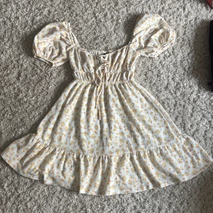 Blommig kortklänning med puffärm - Supersöt kortklänning med volangkant, puffärmar och snörning framtill. Vit botten med små gula och rosa blommor. Gjord i ett lätt, krinklat bomullstyg som känns perfekt för sommaren. Sitter bekvämt med resår i midjan och har en romantisk vibe.