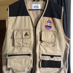 Beige utilityväst från SNS - Utilityväst i beige och svart från SNS med flera fickor framtill, dragkedja i mitten. Västen har meshdetalj på ena fickan och stora fickor även på ryggen.  Endast använd 1 gång. Köpt för 800kr för ett par år sen.