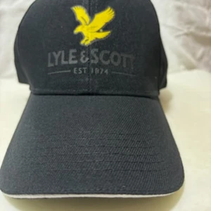 Svart keps från Lyle & Scott - Svart keps från Lyle & Scott med klassisk böjd skärm och justerbar rem baktill. Framtill finns en gul broderad örn och logotypen 'LYLE & SCOTT EST. 1874'. Kepsen är tillverkad i bomull och har ventilerande hål upptill.