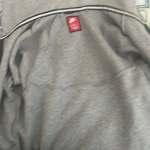 Grå Nike hoodie med dragkedja XL - Snygg grå hoodie från Nike i storlek XL med svarta detaljer och hel dragkedja framtill. Tröjan har huva, långa ärmar och en svart Nike-logga på bröstet. Perfekt för chill eller träning, tillverkad i mjukt material för extra komfort.