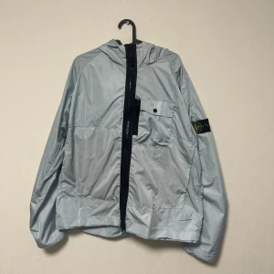 Stone Island Vindjacka - Snygg ljusgrå vindjacka från Stone Island med huva och svart dragkedja. Jackan har en bröstficka med knapp och den klassiska Stone Island-loggan på ärmen. Tillverkad i polyester, perfekt för blåsiga dagar. Oversized passform för en chill vibe.