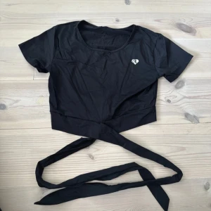 Svart croppad knyt-topp från Womens Best - Svart croppad topp från Womens Best med kort ärm och rund halsringning. Toppen har långa knytband i midjan för en snygg siluett och en liten vit logga på bröstet. Perfekt för dig som gillar sportig och trendig stil.