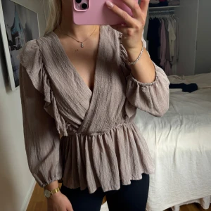 Beige omlottblus med volangdetaljer - Supersöt beige blus med omlottmodell och v-ringning. Toppen har långa ärmar med resår vid handlederna, volangdetaljer på axlarna och ett lätt rynkat, flowy fall nedtill. Perfekt för dig som gillar en romantisk och trendig look.