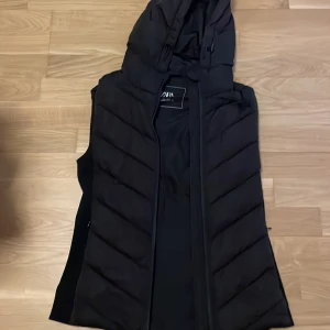 Svart dunväst med huva från Zara - Säljer en svart dunväst från Zara i storlek S. Västen har en stor, vadderad huva med dragsko och snedquiltade paneler. Stängs med dragkedja framtill och är perfekt för lager-på-lager-stil under kyliga dagar. och fickorna är lite tröga 