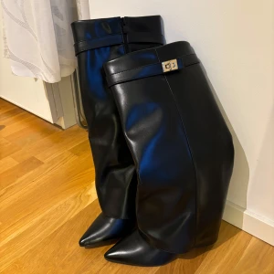 Svarta stövlar med spetsig tå och gulddetalj - Snygga svarta stövlar i skinnimitation med spetsig tå och dekorativt guldfärgat spänne upptill. Stövlarna har en hög och slouchy passform som ger en cool siluett. Perfekta att styla med både kjol och jeans för en trendig look. Aldrig använda. 