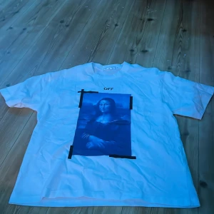 Vit Off-White t-shirt med Mona Lisa-tryck - Vit t-shirt från Off-White med stort blått Mona Lisa-tryck framtill och svarta pilar på ryggen. Klassisk passform och korta ärmar. Snygg streetwear-vibe med unikt grafiskt motiv och tydlig logga. Perfekt för dig som gillar statement-plagg.