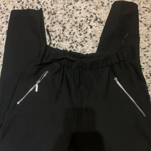 Svarta mjukisbyxor från Miss Selfridge - Svarta mjukisbyxor från Miss Selfridge med dragsko i midjan och två sneda fickor med silvriga dragkedjor. Byxorna har en relaxed fit och är perfekta för en chill och avslappnad stil.