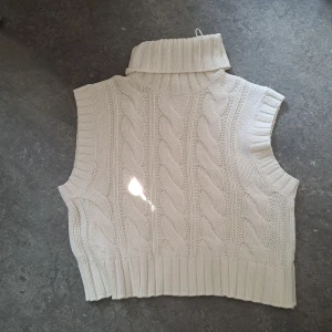 Vit stickad väst med polokrage - Snygg vit stickad väst med polokrage och grovt kabelstickat mönster. Västen är ärmlös och har breda ribbade kanter vid ärmhål och nederkant. Perfekt att slänga över en skjorta eller långärmad tröja för en trendig lager-på-lager-look.