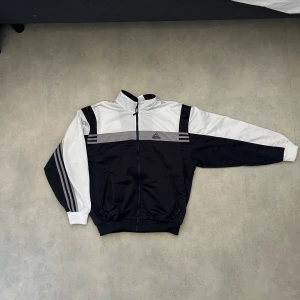 Svart och vit vindjacka från Adidas - Snygg svart och vit vindjacka från Adidas med dragkedja och klassiska ränder på ärmarna. Perfekt för en sportig look. Jackan har en hög krage och två fickor framtill.
