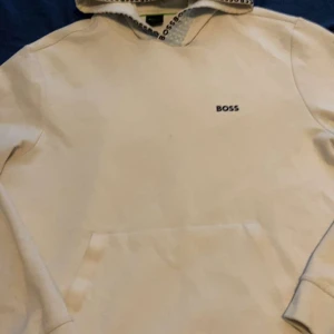 Vit hoodie från Boss - Säljer en stilren vit hoodie från Boss med logotyp på bröstet och mönstrad utsida av huvan. Perfekt för en avslappnad look. 