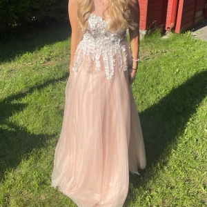 Ljusrosa långklänning med spetsdetaljer - Drömmig ljusrosa långklänning med voluminös tyllkjol och vacker överdel täckt av vita spetsblommor. Klänningen är ärmlös och har en romantisk, flowy siluett som verkligen sticker ut. Perfekt för bal eller student!