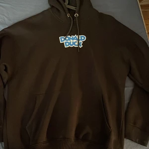 Svart hoodie med Donald Duck-motiv från Nike - Säljer en svart hoodie från Nike med ett coolt Donald Duck-tryck på ryggen. Framsidan har en text med 'Donald Duck' i blått och vitt. Hoodien har en klassisk passform med dragsko i huvan och en stor ficka framtill.