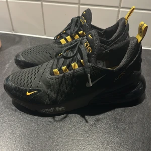 Svarta Nike Air Max med gula detaljer - Snygga svarta Nike Air Max sneakers med gula detaljer och snörning. Skorna har en luftig design med synlig luftdämpning i hälen för extra komfort. Perfekta för både stil och funktion.
