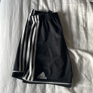 Svarta shorts från Adidas - Snygga svarta shorts från Adidas med de klassiska tre ränderna längs sidorna. Perfekta för sport och träning. Elastisk midja för en bekväm passform.
