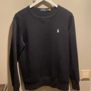 Mörkblå tröja från Polo Ralph Lauren - Säljer en stilren mörkblå tröja från Polo Ralph Lauren med det klassiska logotypbroderiet på bröstet. Tröjan har en rund halsringning och långa ärmar, perfekt för en avslappnad look.