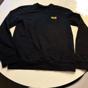 Svart tröja från Emporio Armani storlek M/L - Snygg svart tröja från Emporio Armani med EA7-logga i gult på bröstet. Perfekt för en stilren look. Tröjan har långa ärmar och rund halsringning.