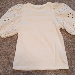 Vit blus med spetsdetaljer - Säljer en söt vit blus med vackra spetsdetaljer på ärmarna. Blusen har en rund hals och korta puffärmar med broderade hålmönster. Perfekt för en stilren och romantisk look. Knapt använd.