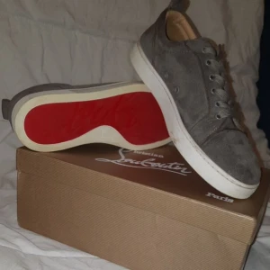 Grå sneakers från Christian Louboutin - Snygga grå sneakers i mocka från Christian Louboutin med den ikoniska röda sulan. Skorna har snörning och en stilren design som passar perfekt för en trendig look.