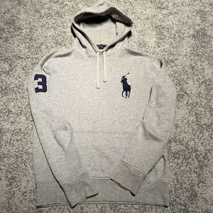 Grå hoodie från Polo Ralph Lauren - Säljer denna sällsynta hoodie från Polo Ralph Lauren med en broderad logga på bröstet och siffran 3 på ärmen. Tröjan har en justerbar huva med snören och en stor ficka framtill. Perfekt för en avslappnad look!
