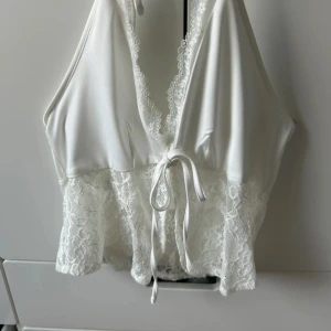 Vit spetsdetaljerad topp - Säljer en vit topp med spetsdetaljer och knytning framtill. Toppen är ärmlös och har en djup v-ringning. Perfekt för en somrig look eller en festlig kväll.